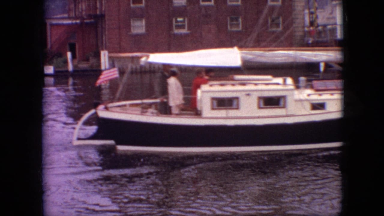 Sailboat with passengers and a small American flag moving near brick waterfront buildings.