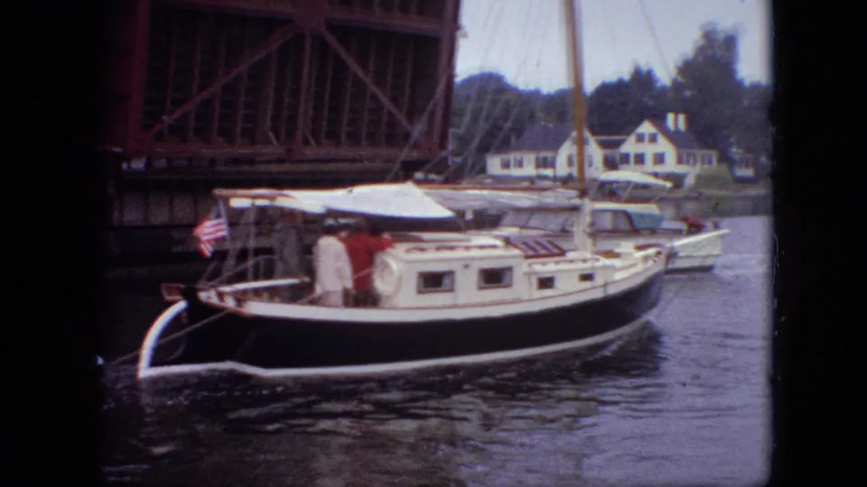 The sailboat continues past the raised drawbridge, showing people on deck, an American flag near the stern, and riverside homes behind it.
