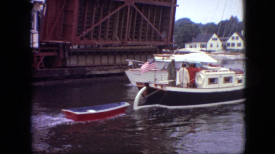 A small red rowboat trails near a white-and-black sailboat as it passes the raised drawbridge, with people aboard and houses across the water.