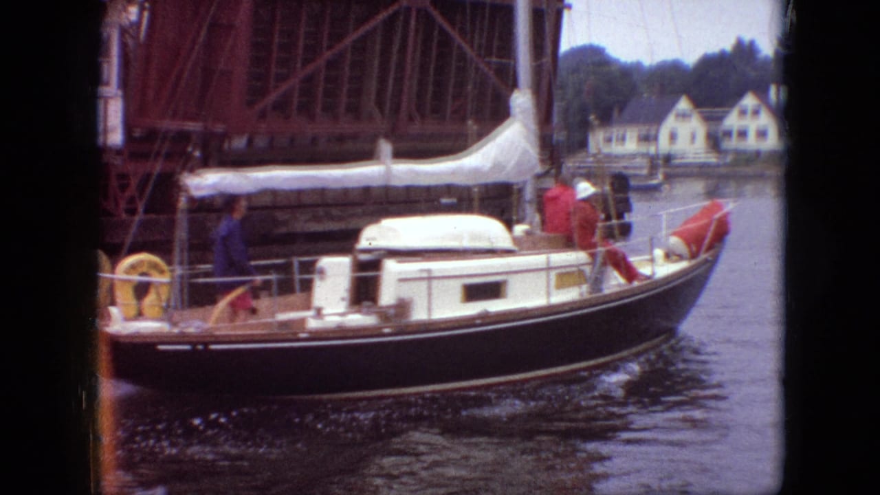 1968 Branson, Missouri home-movie frame showing a dark-hulled cabin sailboat with a lowered white sail moving under a raised bridge, with shoreline houses in the background.