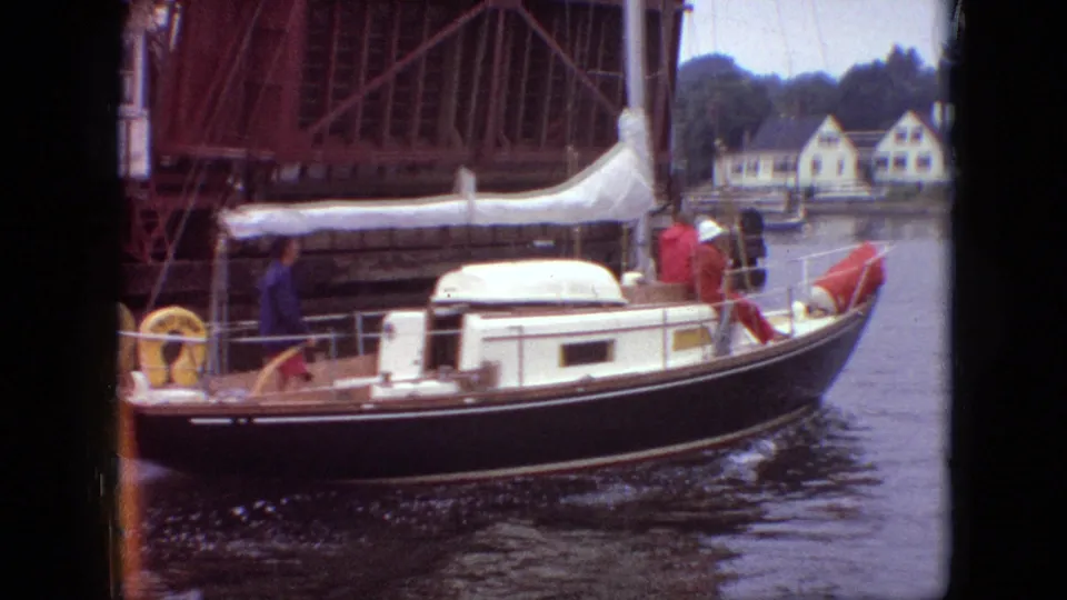 1968 Branson, Missouri home-movie frame showing a dark-hulled cabin sailboat with a lowered white sail moving under a raised bridge, with shoreline houses in the background.