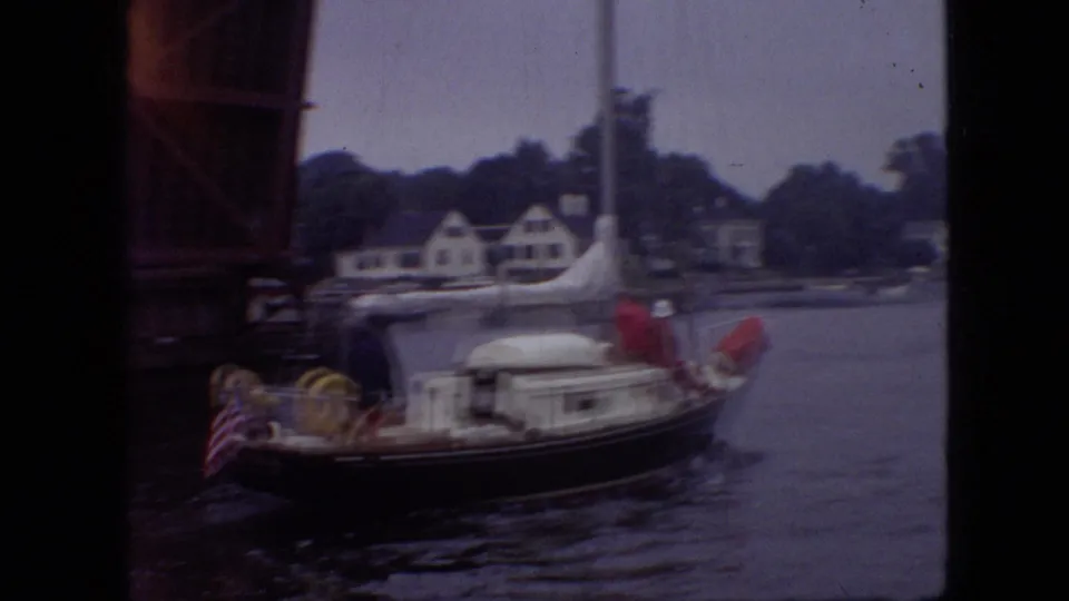 1968 Branson, Missouri home-movie frame showing the cabin sailboat continuing through the bridge area, with an American flag at the stern and houses across the river.
