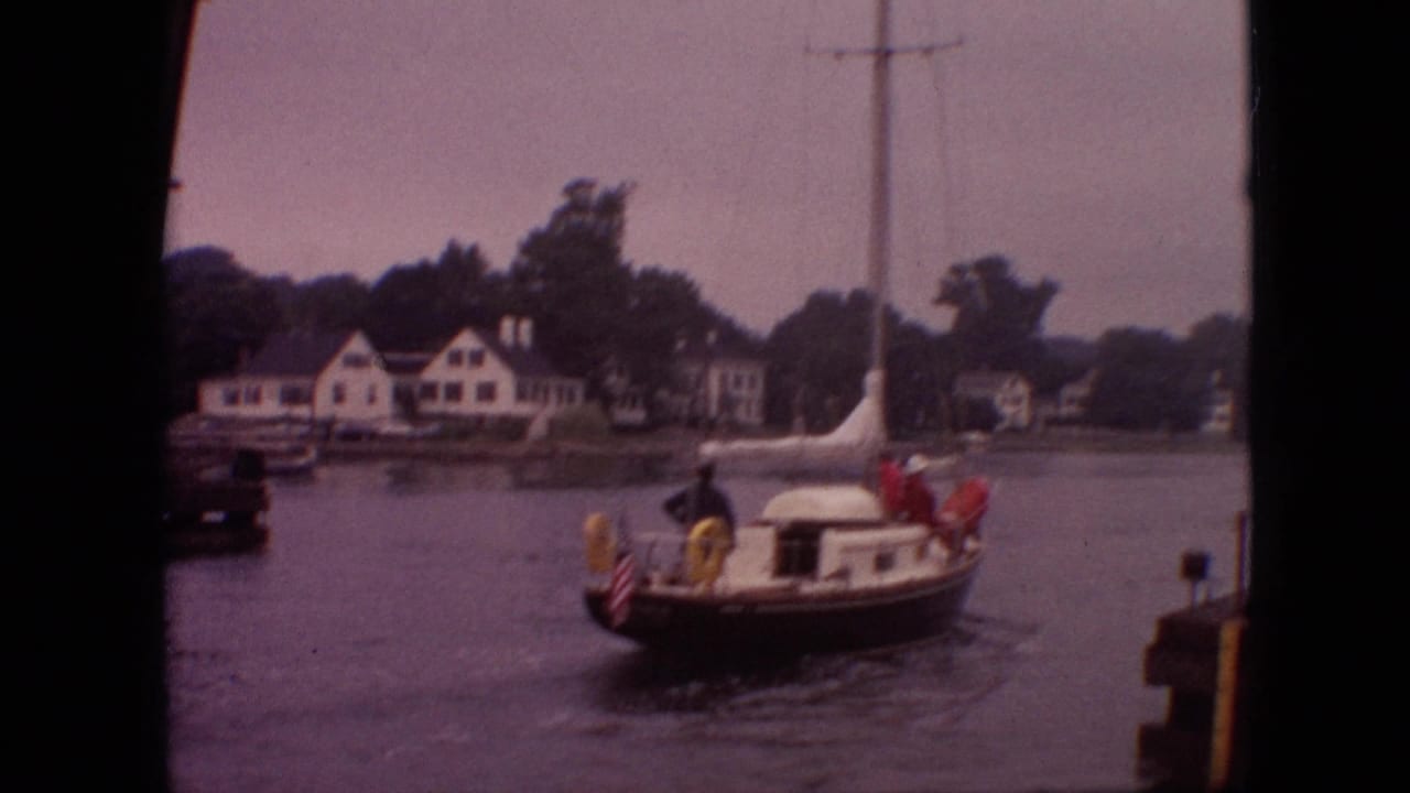 Small sailboat on calm water near shoreline houses and trees.