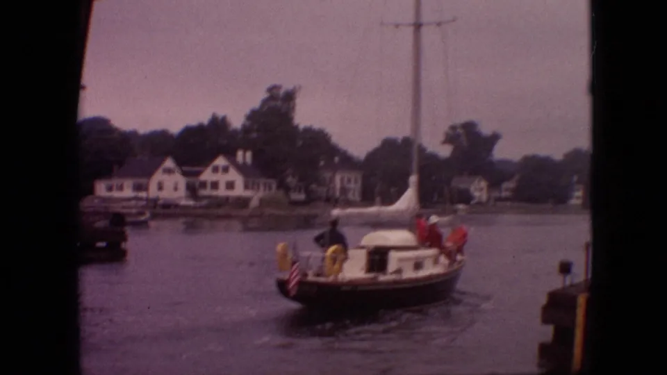 1968 Branson, Missouri harbor view with a small sailboat carrying distant people, shoreline houses, trees, and calm water.