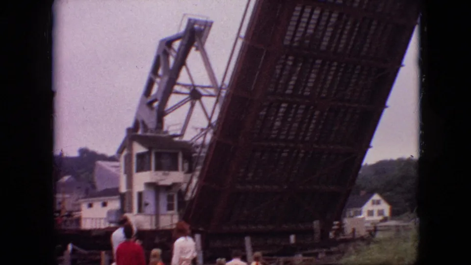 1968 Branson, Missouri frame showing the raised bascule bridge, a small group of people watching near the base, and riverside houses below wooded hills.