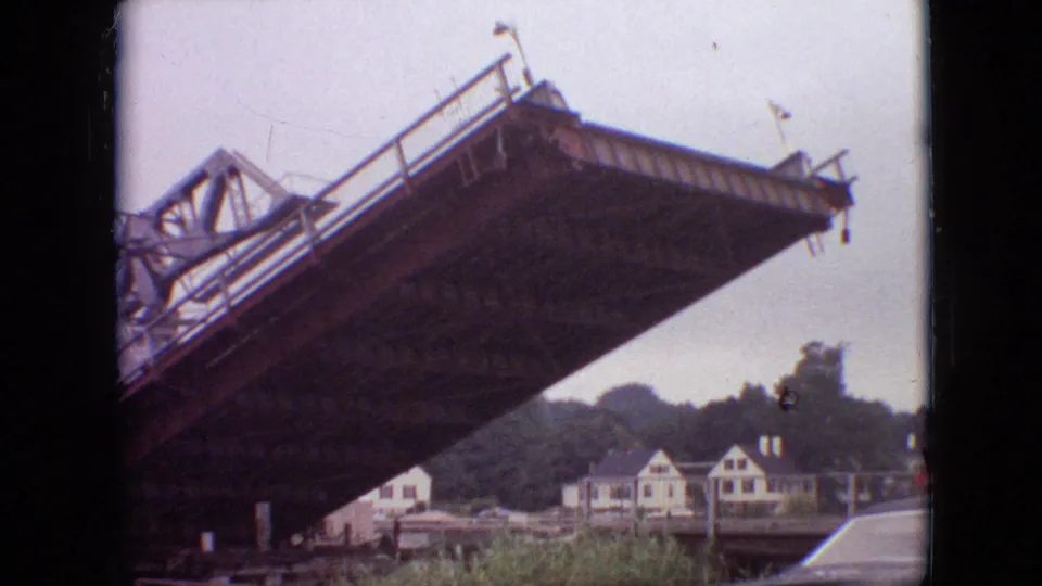 A low-angle 1968 Branson, Missouri 8mm frame shows a large steel drawbridge span still raised, with white riverside houses and trees beyond it.