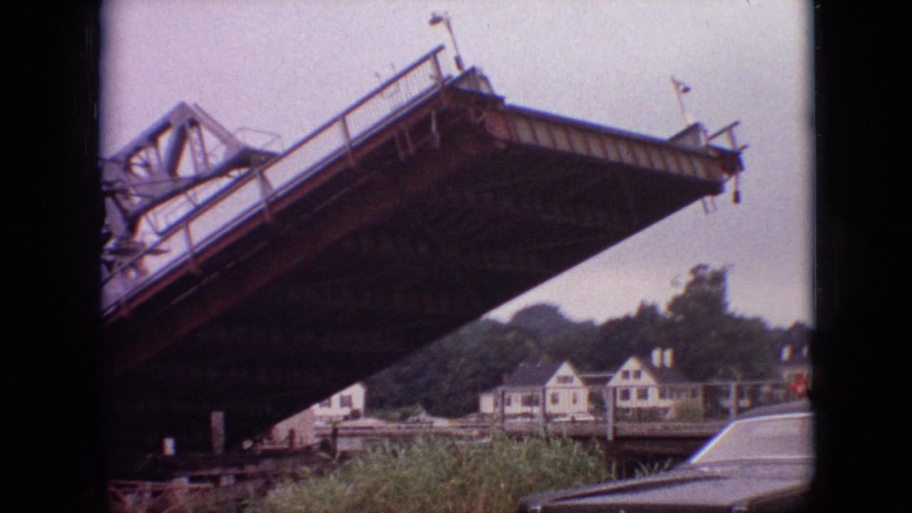 The steel drawbridge appears lower in this 1968 Branson, Missouri frame, with riverside houses, trees, tall grass, and the rear of a parked car in view.