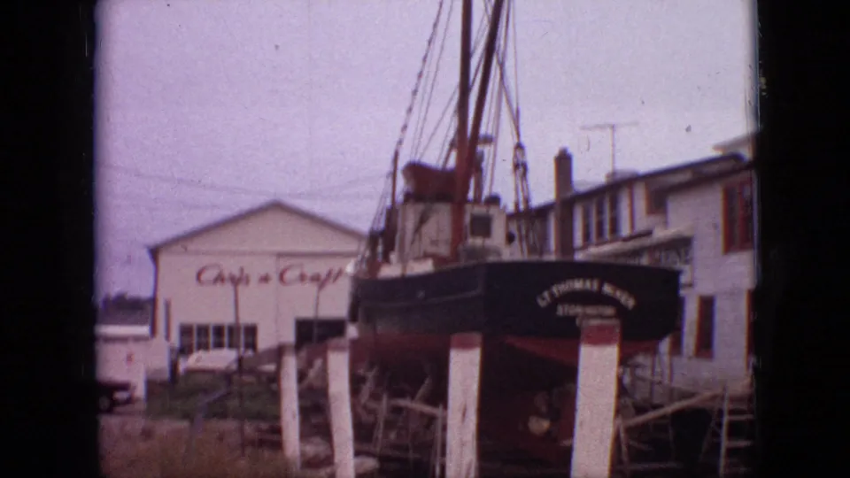 1968 Branson, Missouri 8mm frame with the stern of a dark vessel, readable boatyard signage, support posts, and nearby waterside buildings around the hull.