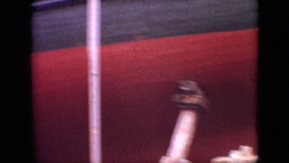 Close 8mm frame of a red-painted hull surface with a pale pipe or rail crossing the foreground and soft, grainy exposure.