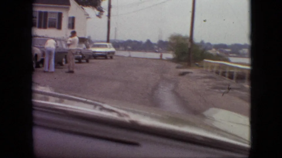 1968 Branson, Missouri roadside view through a windshield, with people near parked cars, a building edge, utility poles, and a railing overlooking lower ground or water.