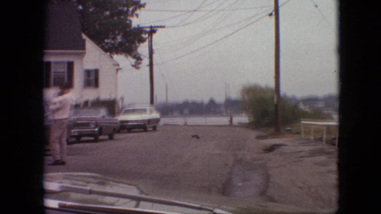 1968 Branson, Missouri home-movie frame from inside a moving car, showing a quiet residential street, parked cars, a white house with dark shutters, overhead wires, and a person in light clothing near the left curb.