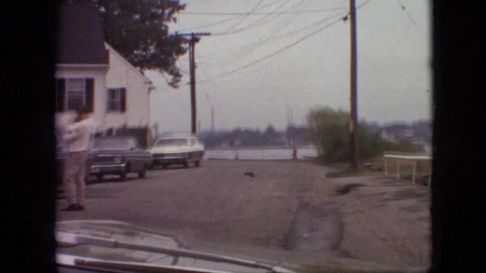 1968 Branson, Missouri home-movie frame from inside a moving car, showing a quiet residential street, parked cars, a white house with dark shutters, overhead wires, and a person in light clothing near the left curb.