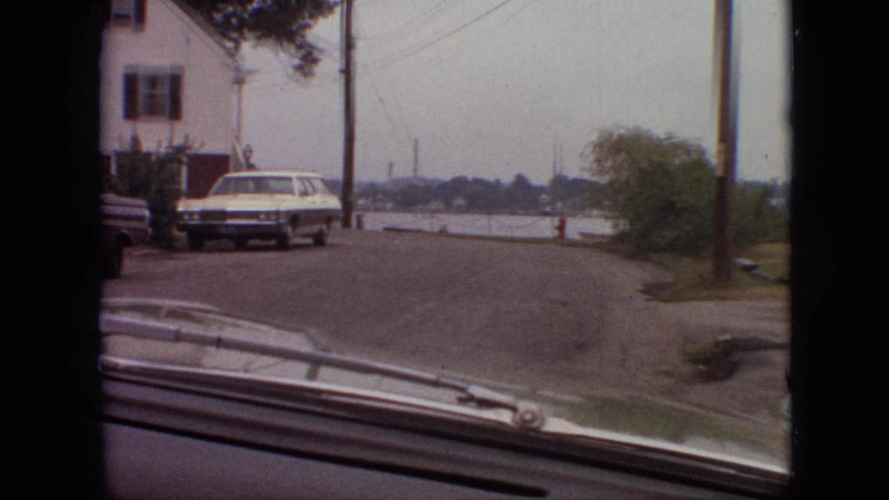 View over a car hood toward a white station wagon, house, utility poles, water, and a red fire hydrant.