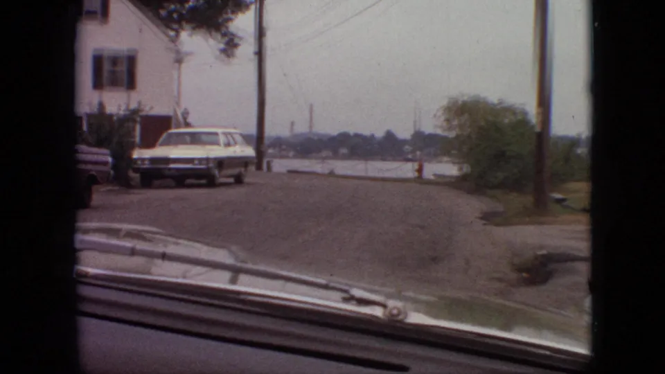 1968 Branson, Missouri driving frame showing the camera car approaching a bend or end of a residential street with a white station wagon, house, utility poles, water beyond the road, and a red fire hydrant.