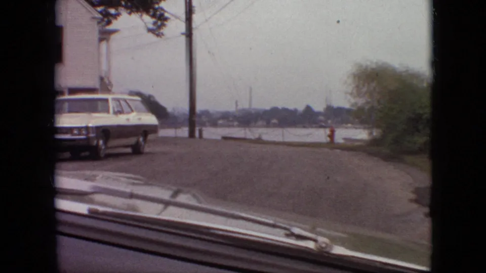 1968 Branson, Missouri home-movie frame from a moving vehicle, with a white station wagon on the left, a residential street ahead, utility poles, a red hydrant, and water with distant industrial-looking stacks on the horizon.