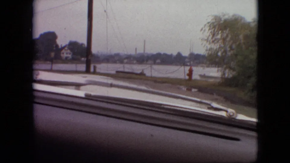 1968 Branson, Missouri driving frame looking past the car hood and windshield wipers toward a utility pole, red fire hydrant, shoreline trees, water, and faint distant structures across the water.