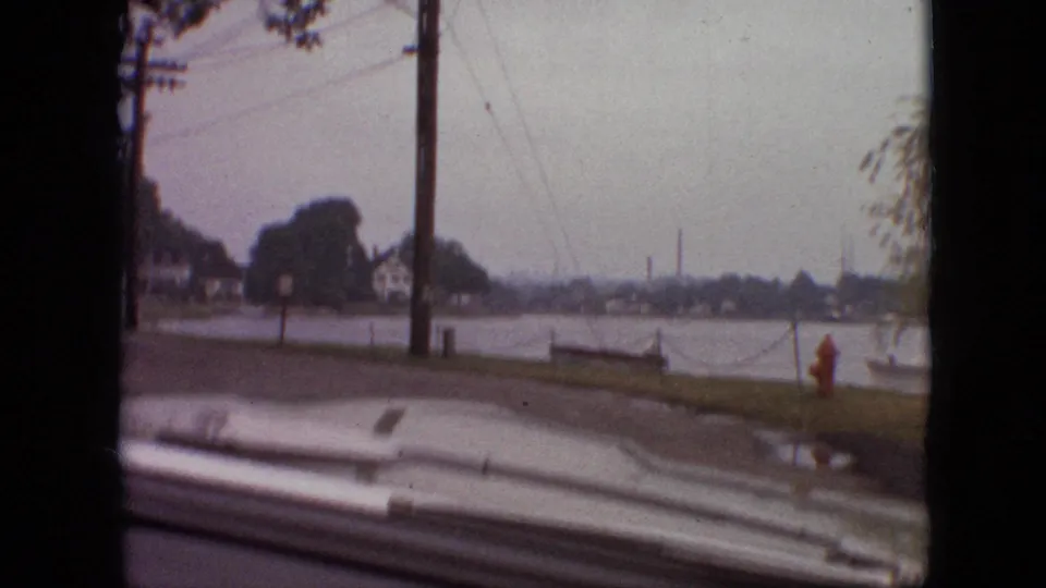 1968 Branson, Missouri 8mm frame from a moving car showing a lakeside residential road, utility poles, houses, a red fire hydrant, and the car hood at the bottom edge.