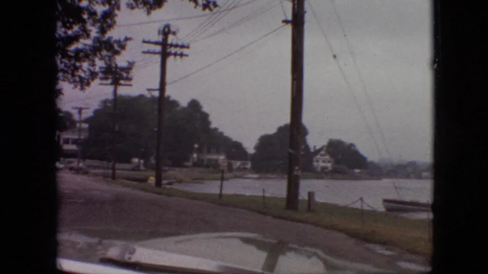 1968 Branson, Missouri home-movie frame showing a quiet road along the lake with wooden utility poles, homes among trees, a small boat near shore, and the car hood visible.