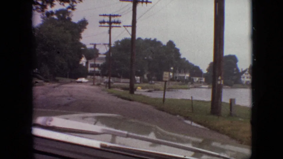 1968 Branson, Missouri 8mm frame from a moving vehicle showing a curving lakeside neighborhood road, utility poles, houses, trees, roadside signs, and windshield reflections.