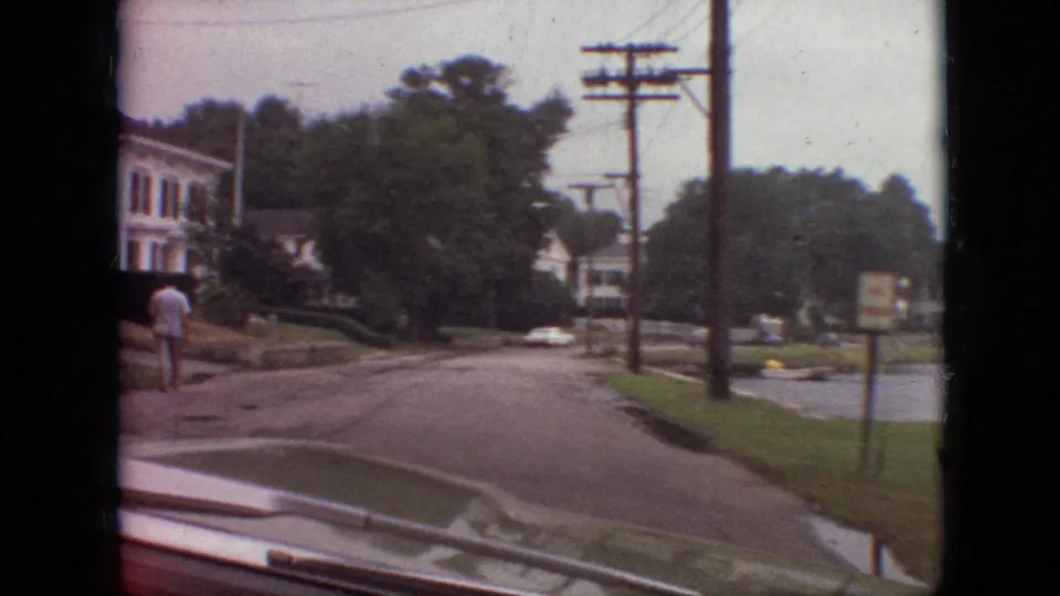 1968 Branson, Missouri home-movie frame showing the car approaching a lakeside residential stretch with houses, trees, utility poles, and a distant pedestrian walking near the sidewalk.