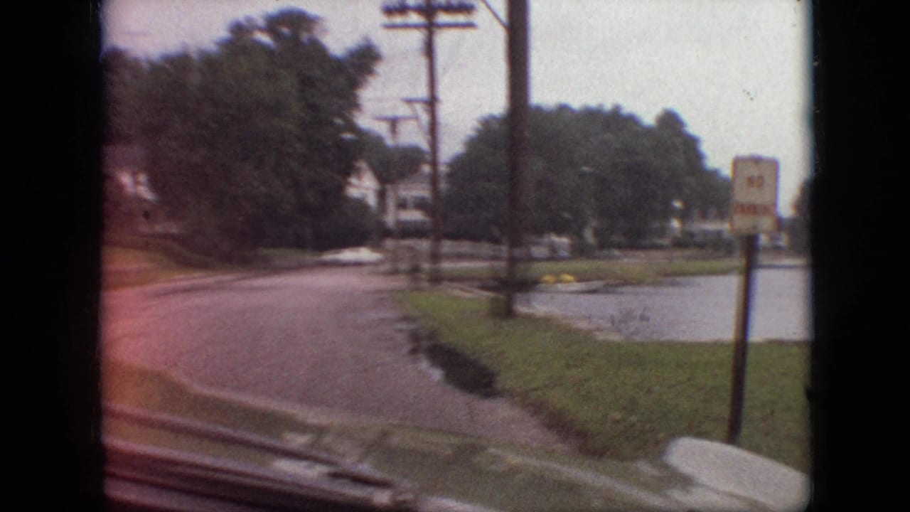 1968 Branson, Missouri home-movie frame from a moving vehicle showing a lakeside road, grass verge, trees, utility poles, and a No Parking sign near the water.