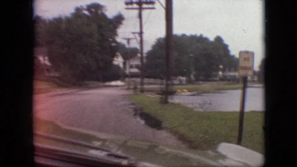 1968 Branson, Missouri home-movie frame from a moving vehicle showing a lakeside road, grass verge, trees, utility poles, and a No Parking sign near the water.