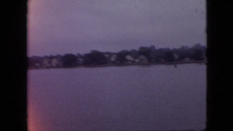 1968 Branson, Missouri home-movie frame showing calm lake water, a small boat, and a distant tree-lined shoreline with houses under a cloudy sky.