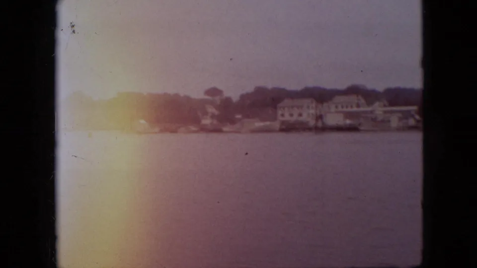1968 Branson, Missouri home-movie frame with lake water in the foreground, houses and trees across the shoreline, and a warm light flare along the left side of the film frame.