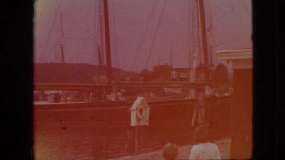 1968 Branson, Missouri waterfront frame with a large docked sailboat, tall rigging, a small dockside structure, and partial people near the edge of the shot.