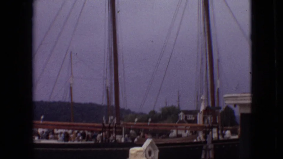 1968 Branson, Missouri home-movie frame showing people gathered along a dock near sailboat masts, railings, and a low tree-lined shoreline.