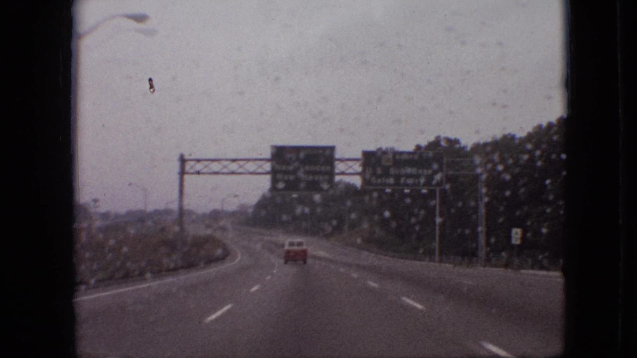 1968 8mm home-movie view through a wet windshield, showing a lightly trafficked highway with a red car ahead and green directional signs under an overcast sky.