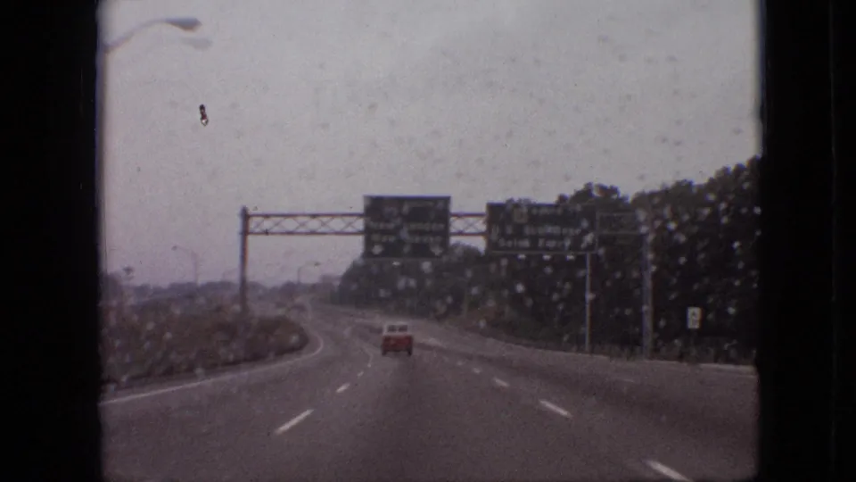 1968 8mm home-movie view through a wet windshield, showing a lightly trafficked highway with a red car ahead and green directional signs under an overcast sky.