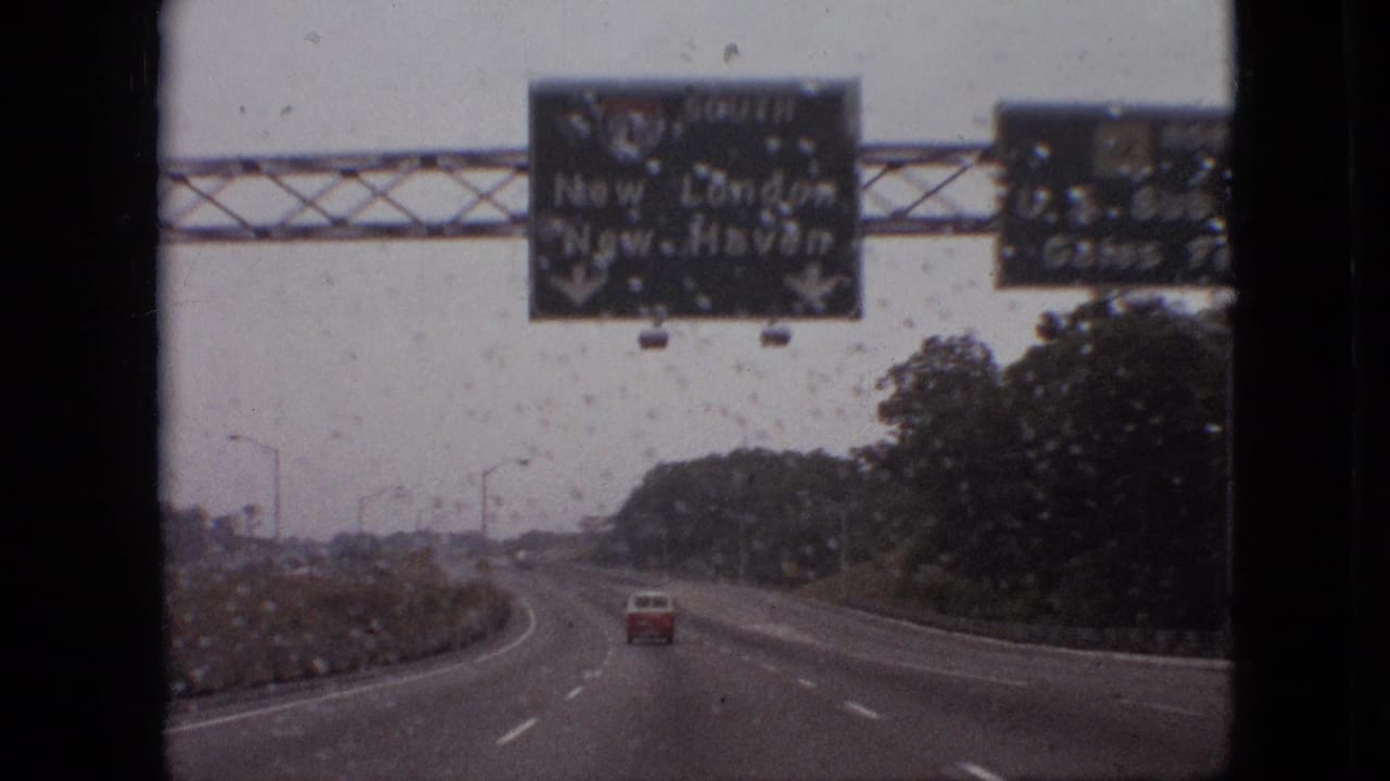 A closer 1968 8mm road view shows rain on the windshield, green overhead guide signs, a red car ahead, and a curving highway bordered by trees.
