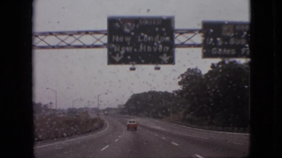 A closer 1968 8mm road view shows rain on the windshield, green overhead guide signs, a red car ahead, and a curving highway bordered by trees.