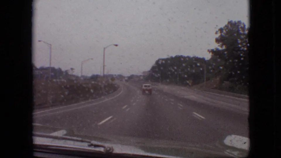 The home-movie camera looks through a rain-marked windshield at an open multi-lane highway, a distant vehicle, roadside trees, and the lower edge of the car interior.