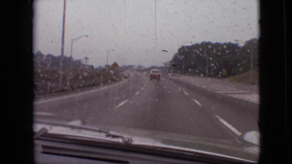 A rain-dotted 8mm frame shows the highway continuing ahead with a red vehicle in the lane, trees at the roadside, streetlights, and the filming car hood visible at the bottom.