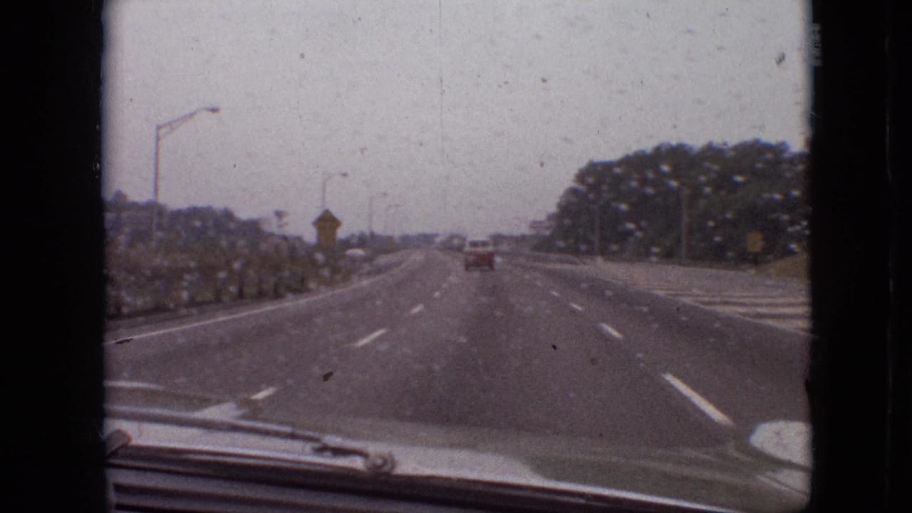 1968 Branson, Missouri home-movie view through a rain-speckled windshield, showing a broad highway, roadside plantings, distant traffic, and the car hood at the bottom of frame.