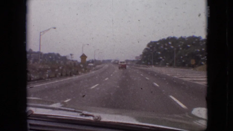 1968 Branson, Missouri home-movie view through a rain-speckled windshield, showing a broad highway, roadside plantings, distant traffic, and the car hood at the bottom of frame.