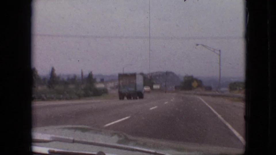 Rainy 1968 Branson road scene filmed from inside a car, showing a semi-truck ahead, lane markings, roadside signs, and hazy hills beyond the highway.