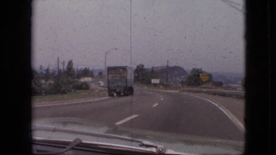 Final frame of the 1968 Branson highway drive, with the camera looking past the car hood toward a semi-truck, roadside signs, trees, and a hazy wet roadway.