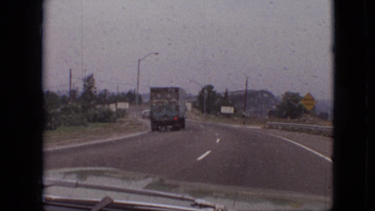 1968 Branson, Missouri home-movie frame showing a moving-car view of a truck ahead on a curving roadway with roadside vegetation, utility poles, and distant hills.