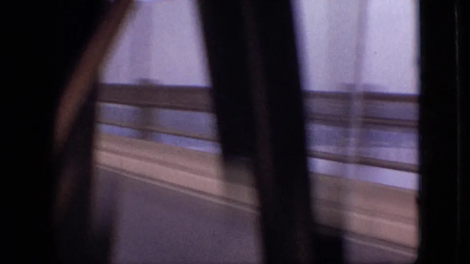 Passenger-side 8mm view during the bridge crossing, with a dark interior edge in the foreground, blurred railings, road surface, and water glimpsed beyond.