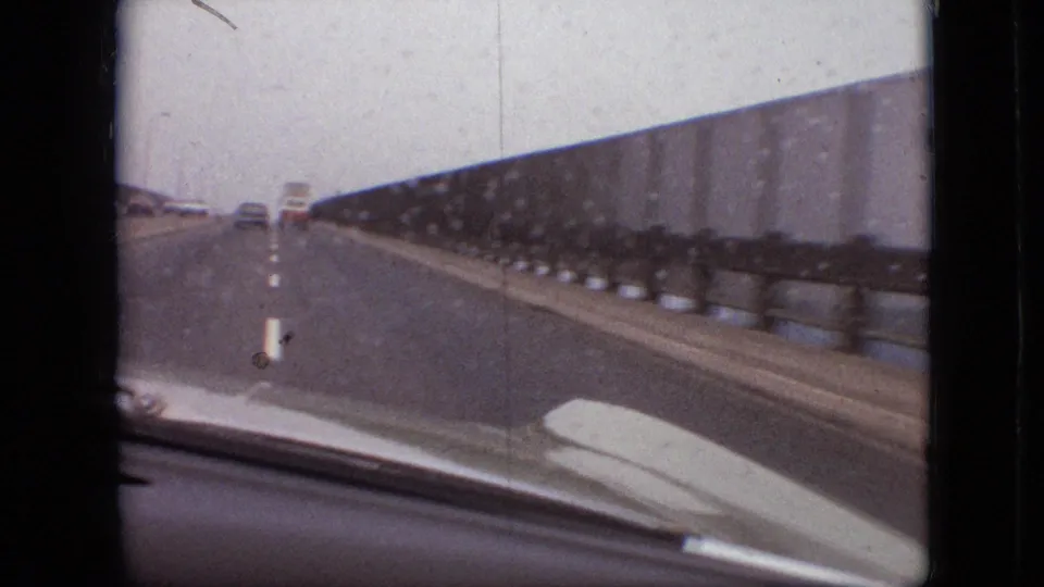 Forward view from the moving car in the 1968 Branson, Missouri footage, showing vehicles ahead, dashed lane markings, a concrete bridge barrier, and the car hood at the bottom edge.