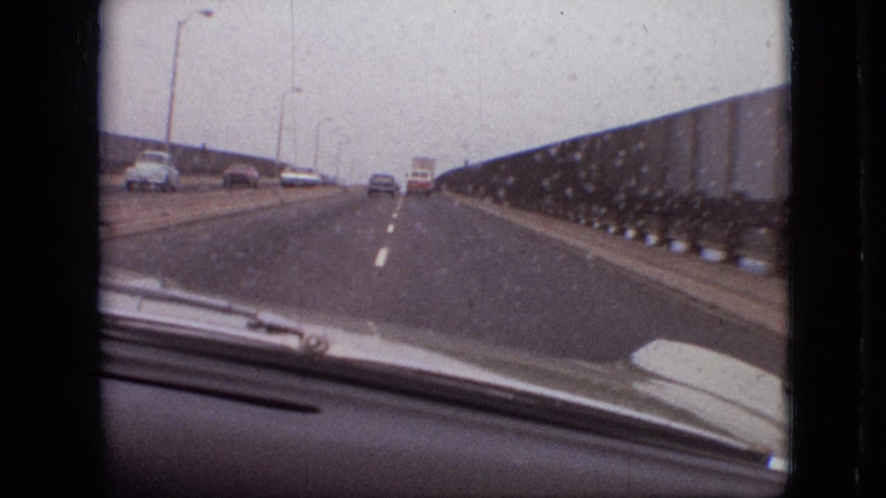 1968 Branson, Missouri home-movie frame looking through a rain-speckled windshield as a car drives across a multi-lane bridge behind sedans and a white van.