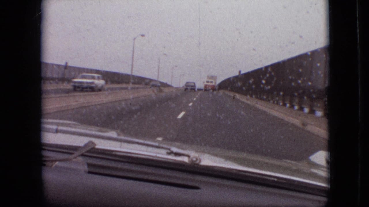 Forward-facing 8mm view through a rain-marked windshield on a Branson, Missouri bridge in 1968, with a sedan and white van visible on the road ahead.