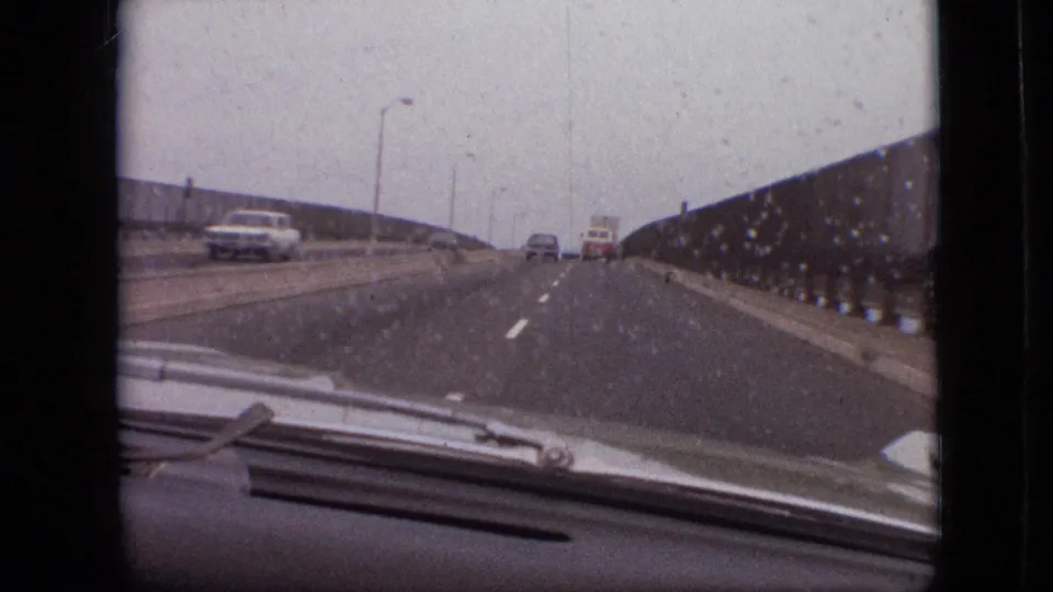 Forward-facing 8mm view through a rain-marked windshield on a Branson, Missouri bridge in 1968, with a sedan and white van visible on the road ahead.