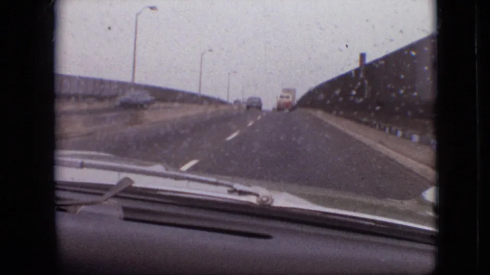 Home-movie frame from inside a moving car on a rainy Branson, Missouri bridge, showing the windshield wiper, dashboard, roadway, bridge wall, and vehicles farther ahead.
