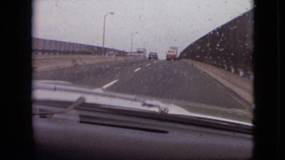 1968 8mm travel frame in Branson, Missouri showing a rain-speckled windshield view toward a white van and car on a multi-lane bridge under gray sky.