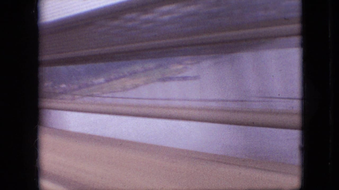 1968 Branson, Missouri home-movie frame showing a soft, blurred view through horizontal window slats toward roadway or guardrail bands and distant water.