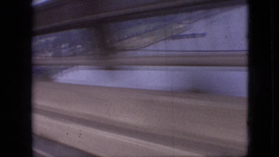 1968 Branson, Missouri home-movie still showing a slatted, motion-blurred view of pale water, shoreline, and broad tan roadway or guardrail shapes.